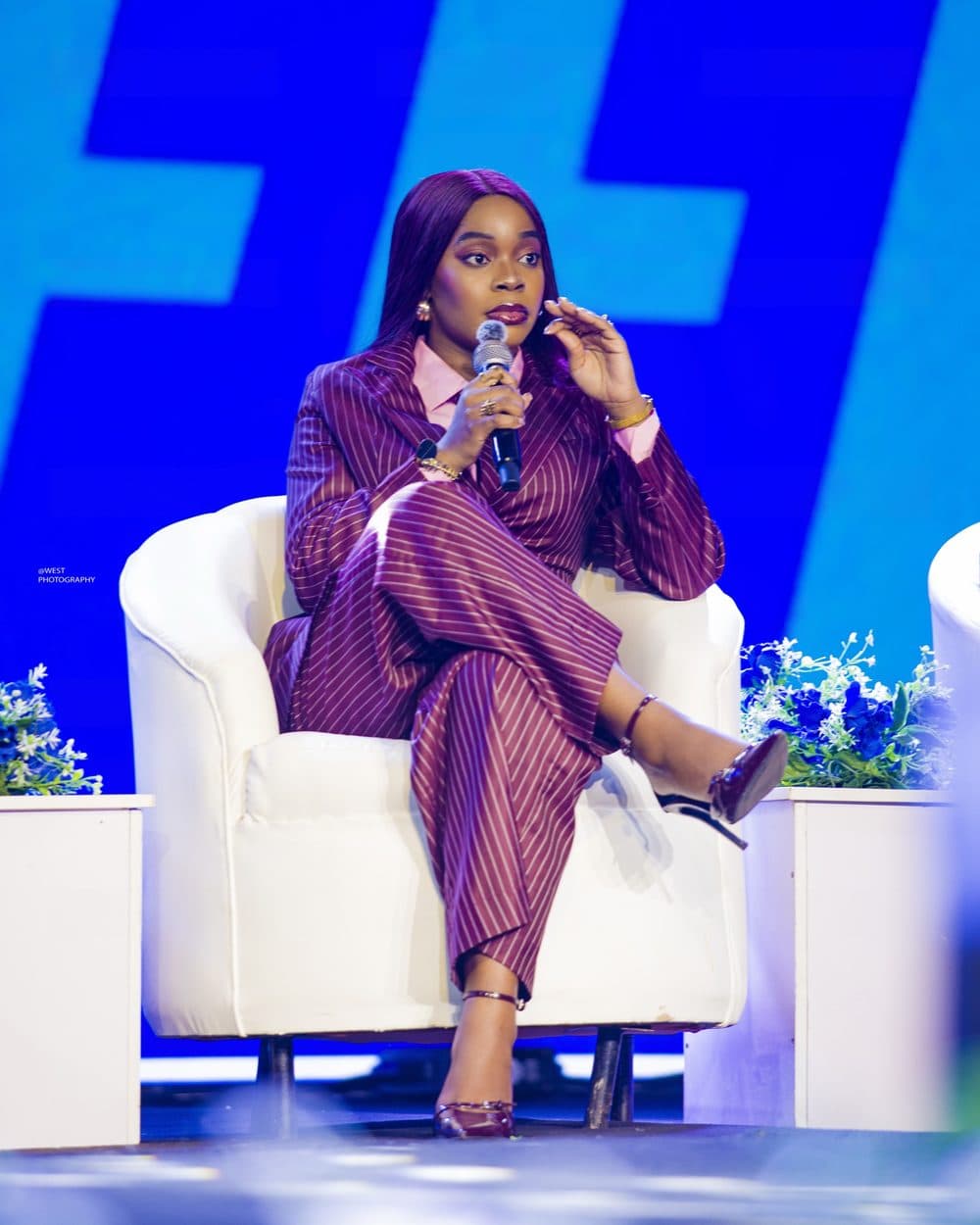 Achie speaking on stage during a panel discussion, seated in a white armchair